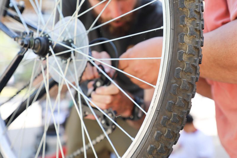 Sur cette photo, on voit une scène en gros plan où deux personnes sont en train de réparer la roue d'un vélo. L’image est prise à travers les rayons de la roue, qui occupent une grande partie du premier plan. Les mains des deux personnes sont visibles, concentrées sur un composant situé près de la chaîne et du dérailleur. L’une des personnes porte un t-shirt foncé, l’autre un vêtement rose orangé. Le pneu visible est large, typique des vélos tout-terrain. À l’arrière-plan, flou, on distingue d'autres personnes et une ambiance extérieure, lors d’un atelier communautaire. L'image donne une impression de collaboration et de concentration.