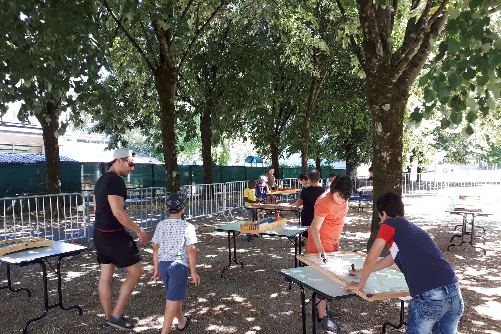 Sous les arbres, des jeunes font des jeux sur des tables.