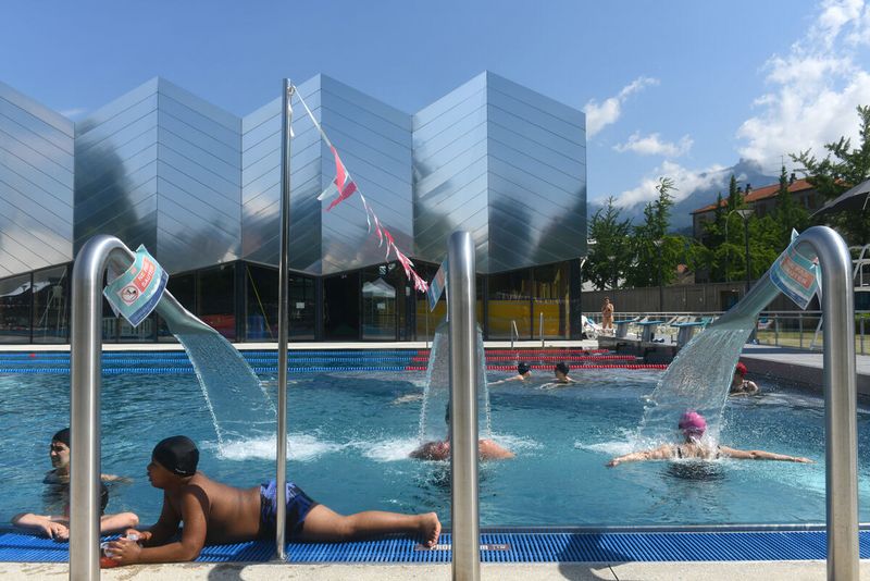 Au premier plan, des personnes profitent des jets d'eau du grand bassin extérieur. Au second plan, on observe des personnes nager. Enfin au dernier plan, on peut admirer l'architecture du bâtiment composée sur sa partie supérieure de chevrons métalliques faisant penser à des vagues. 