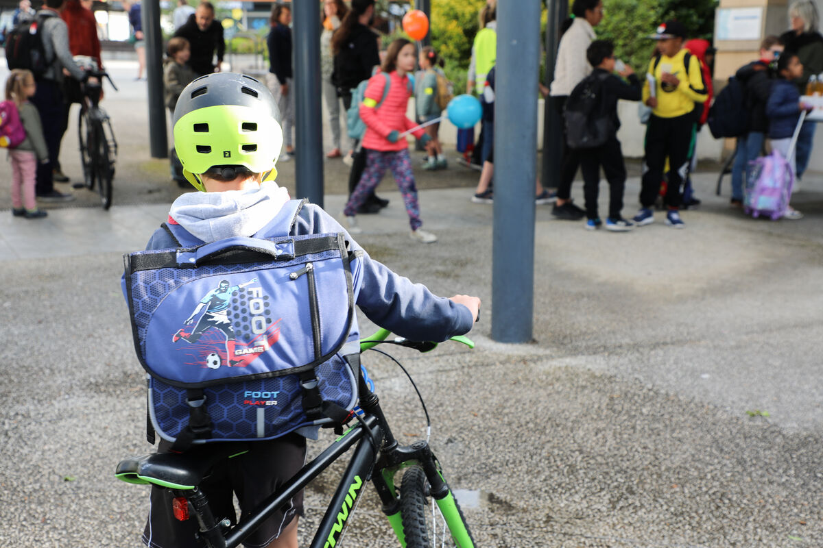 On voit un jeune cycliste de dos avec un sac à dos sur les épaules, un casque sur la tête et son vélo à la main. Il marche en direction de son école.