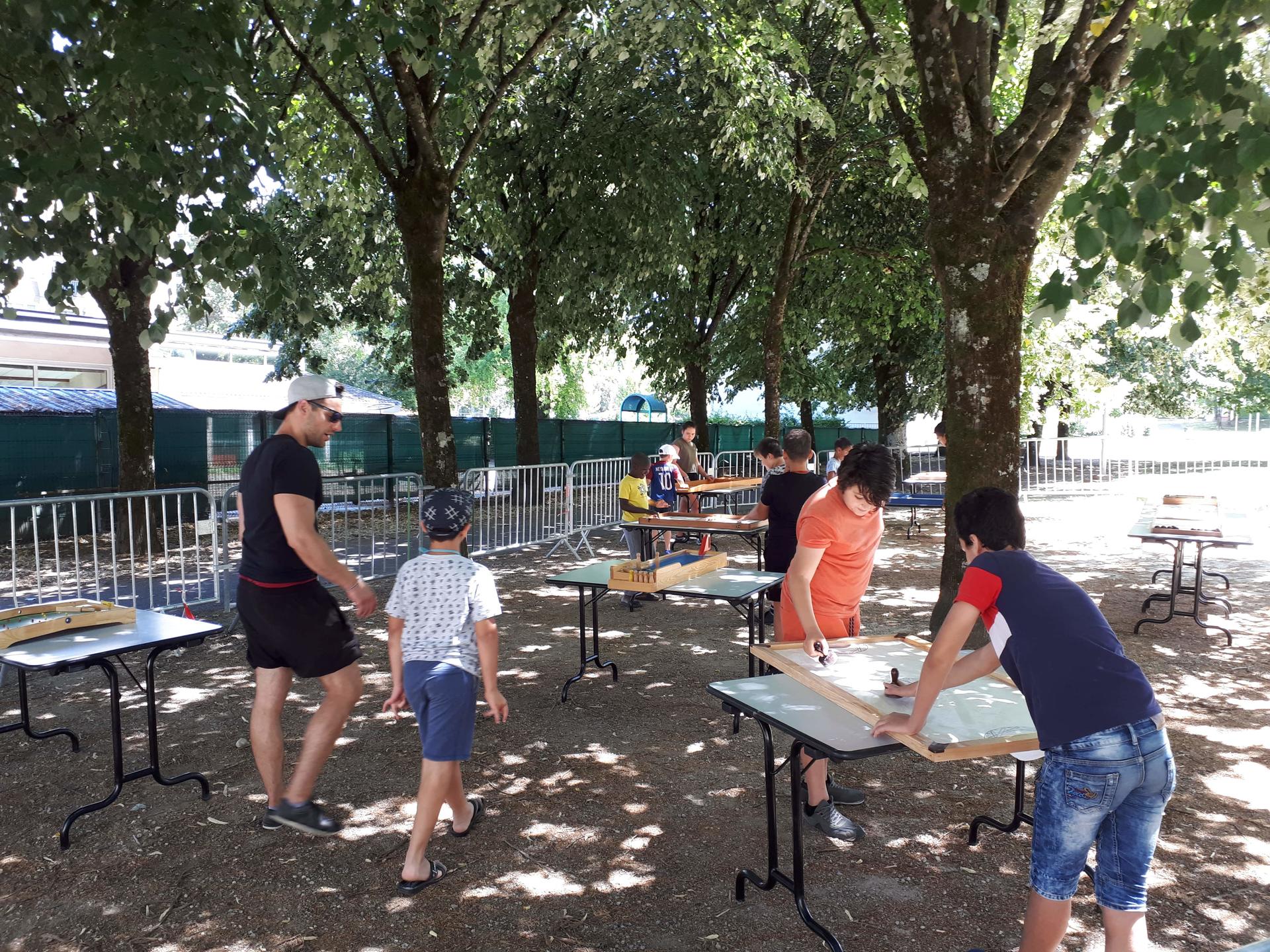 Sous les arbres, des jeunes font des jeux sur des tables.