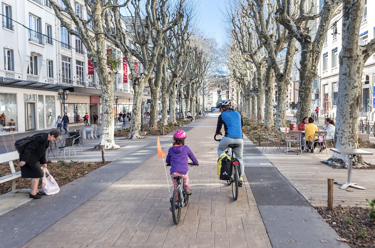 Une large rue piétonne bordée d’arbres sans feuilles. Au centre, un adulte et un enfant roulent tranquillement à vélo sur une voie bien aménagée. L’enfant porte un casque rose. Des passants marchent sur les trottoirs, d’autres sont assis à des terrasses de café. Les bâtiments sont blancs, hauts de plusieurs étages, avec des vitrines de magasins au rez-de-chaussée. L’ambiance est calme et lumineuse, comme un jour de début de printemps.