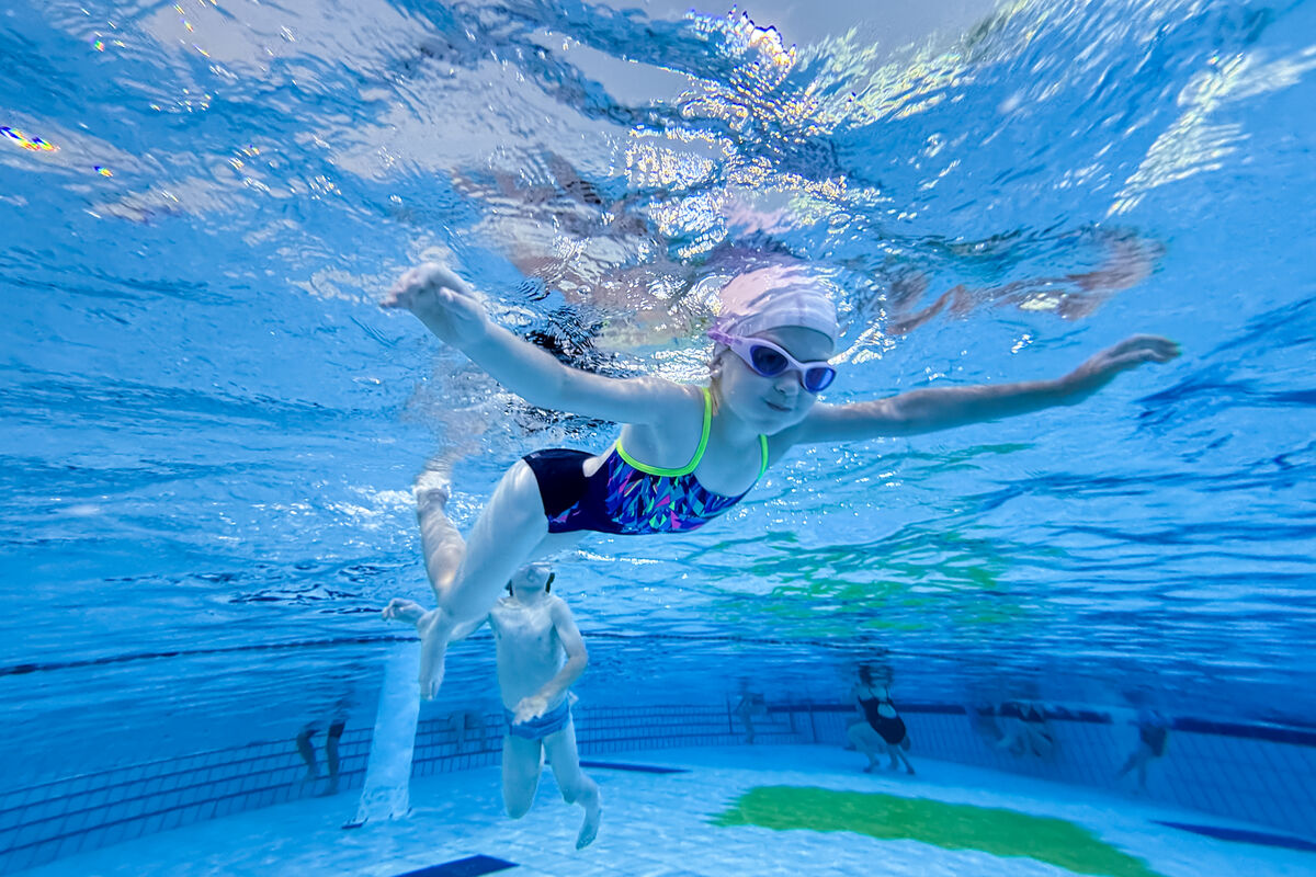 Une enfants nage sous l'eau. Elle porte des lunettes de nage et un maillot bleu foncé.