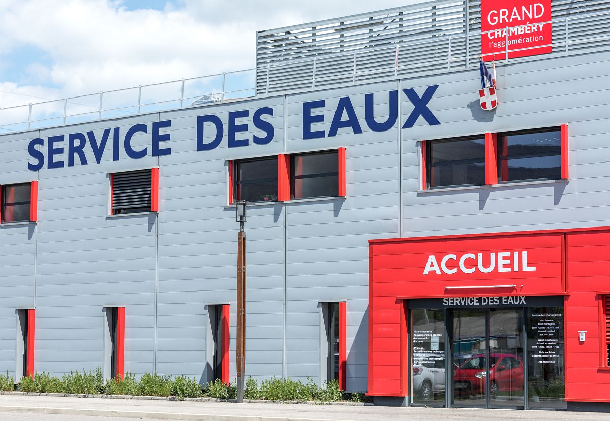 Un grand bâtiment gris avec le logo de Grand Chambéry, un panneau "service des eaux" et en rouge une porte coulissante avec écrit dessus "Accueil".