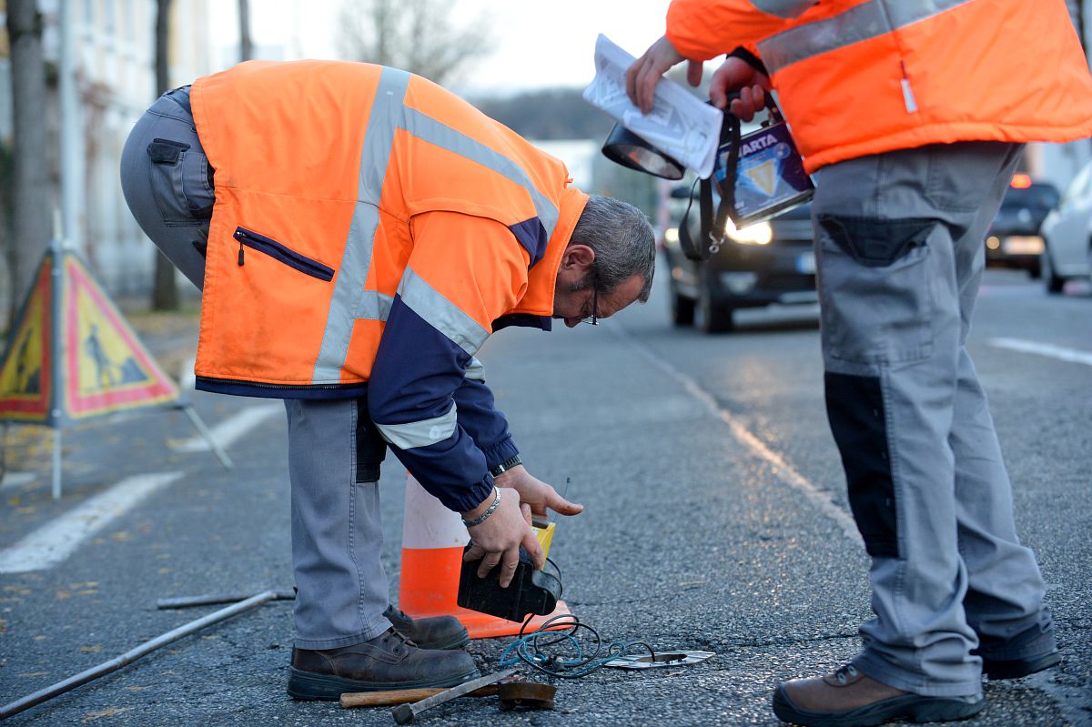 Deux agents avec des vestes oranges de chantier sur une route. L'un des agents a un petit appareil dans la main et il descend un capteur dans le trou de la route.