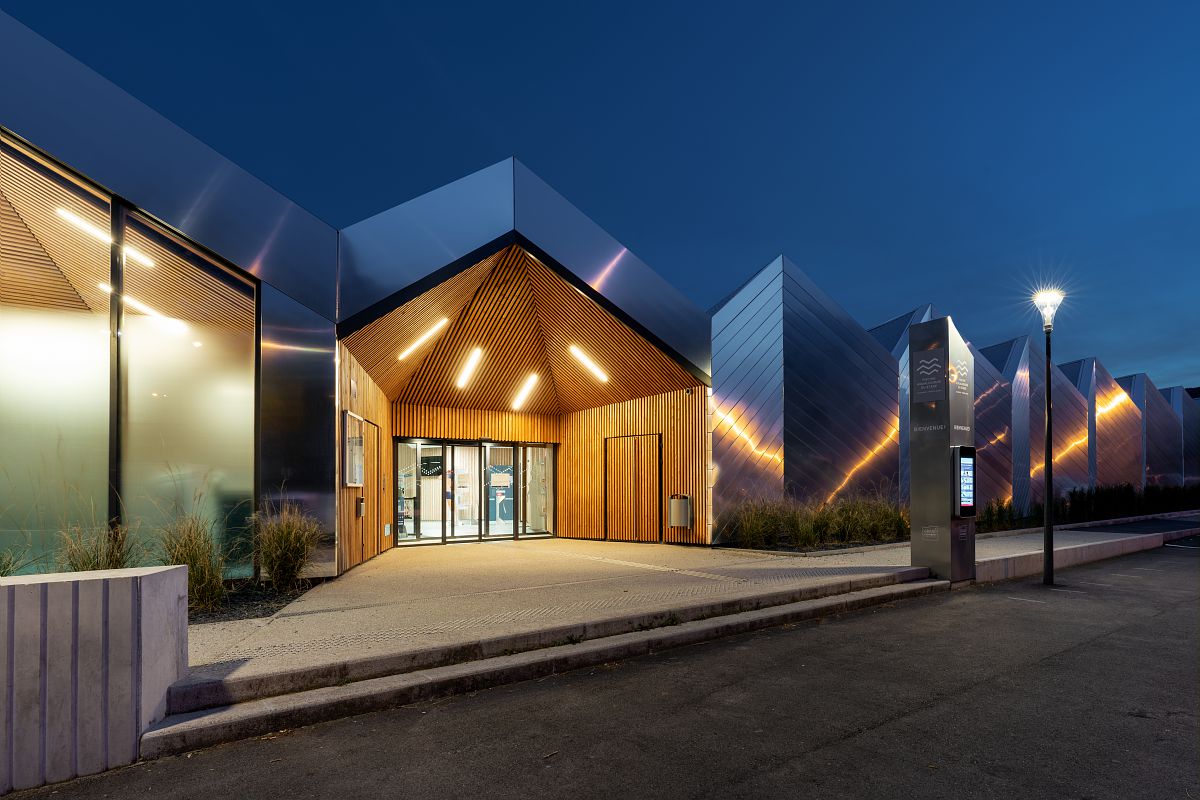 Photo prise de nuit, on perçoit l'entrée éclairée de la piscine aqualudique du Stade. La lumière éclaire l'architecture composée de chevrons métalliques en guise de toit. Le tout fait penser au mouvement de l'eau. 