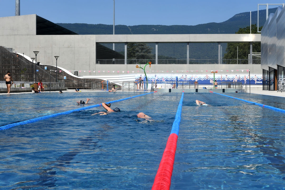 Vue d'ensemble du bassin extérieur et de ses nageurs qui profitent de la piscine mais également des jeux d'eau et des toboggans. On aperçoit en arrière plan le bâtiment du stade de rugby collé aux toboggans et bien plus loin une vue sur la montagne.